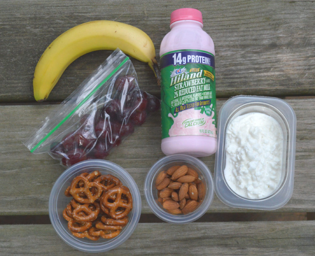 bananas, grapes in bag, Hiland dairy strawberry milk, pretzels, almonds and cottage cheese on a picnic table