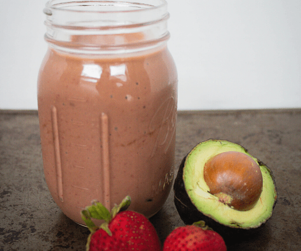 Chocolate strawberry and avocado smoothie in glass jar with 2 fresh strawberries and 1/2 avocado next to it