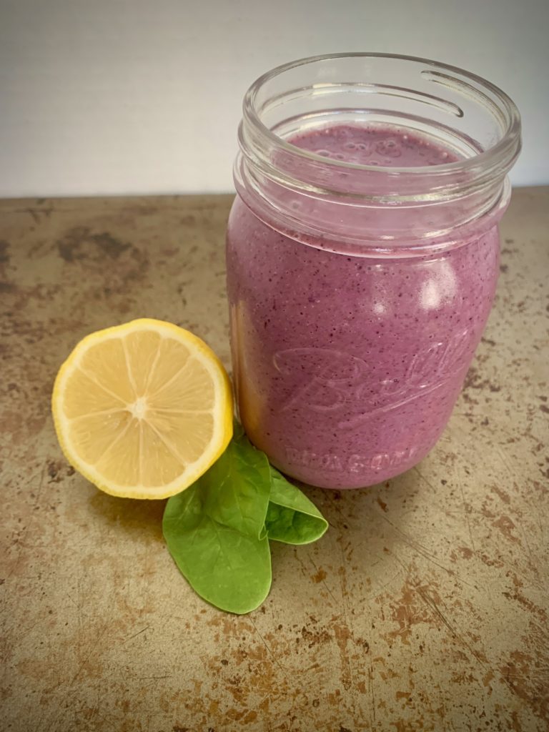 Light purple berry lemon smoothie with vegetables in glass jar next to sliced lemon and fresh spinach