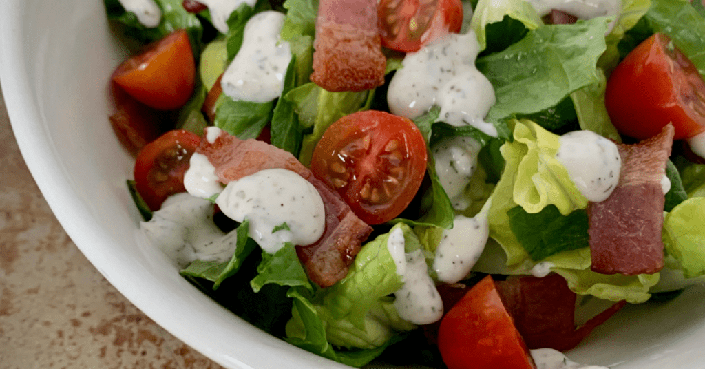 Romaine lettuce with diced bacon, grape tomatoes and ranch dressing in a white bowl. 