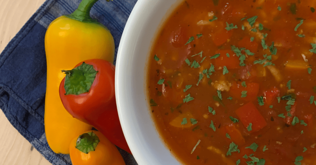 Stuffed pepper soup in white bowl next to three fresh bell peppers.