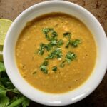 White bowl with lentil curry soup next to lime wedges and cilantro.