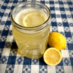 Glass of lemonade in ball jar on blue gingham tablecloth next to whole lemon and 1/2 cut lemon