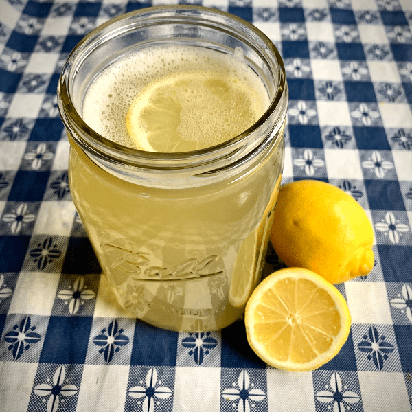 Glass of lemonade in ball jar on blue gingham tablecloth next to whole lemon and 1/2 cut lemon