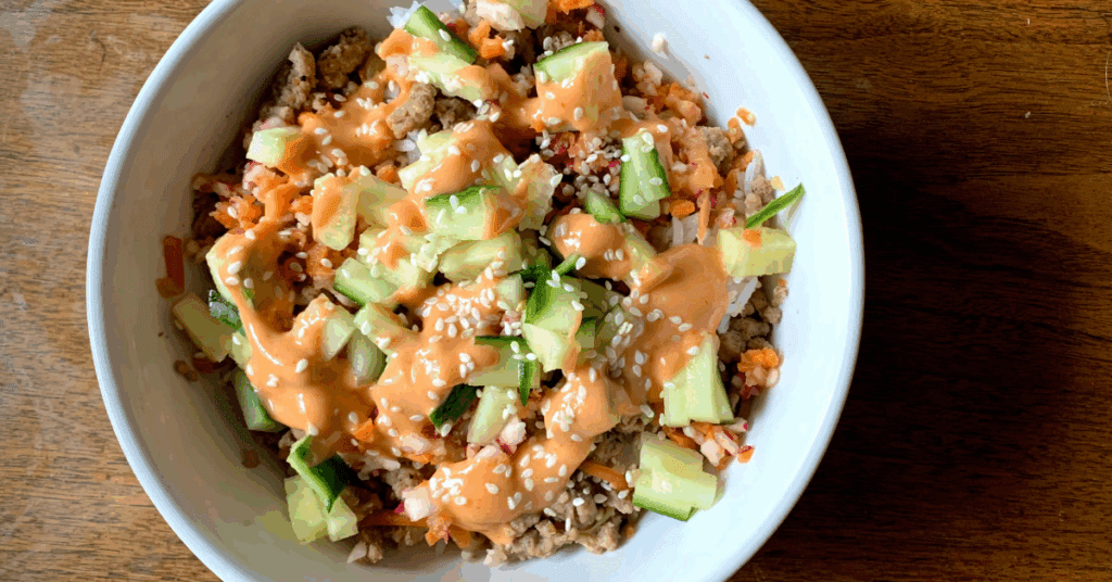 White bowl with bahn mi - Rice, topped with ground pork, diced cucumbers and carrots drizzled with spicy mayo and topped with sesame seeds