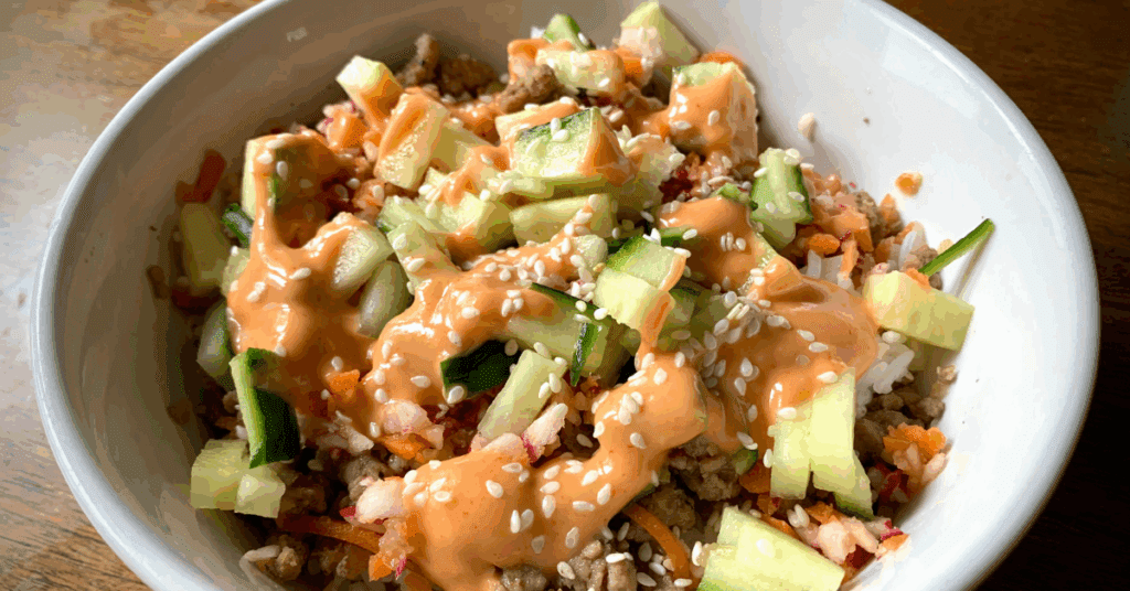 White bowl with bahn mi - Rice, topped with ground pork, diced cucumbers and carrots drizzled with spicy mayo and topped with sesame seeds