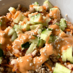 White bowl with bahn mi - Rice, topped with ground pork, diced cucumbers and carrots drizzled with spicy mayo and topped with sesame seeds